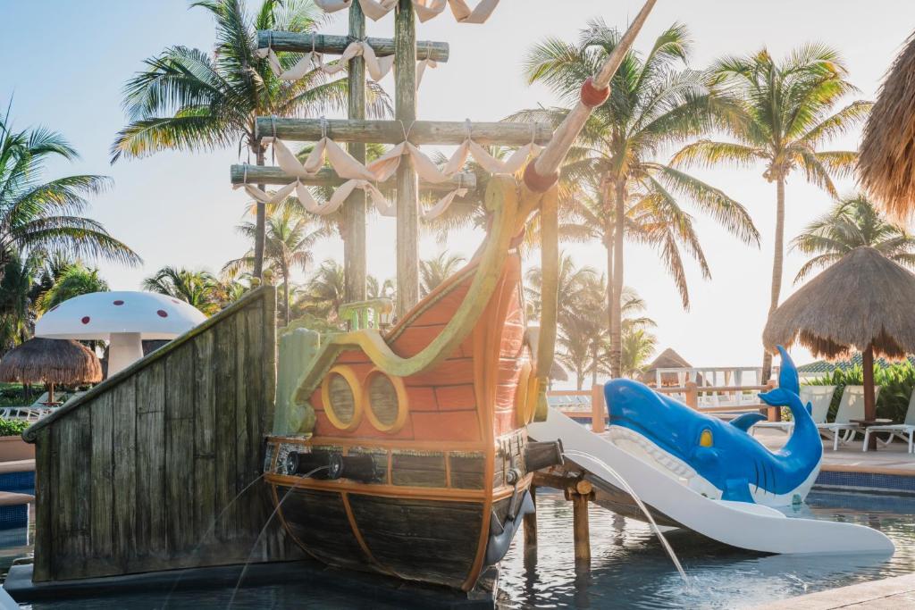 Krāsains ūdens slidkalniņš ar kuģa dizainu un zilu vaļu fomu, izvietots pie baseina tropiskā vidē ar palmām un saulainu fonu. Ideāla vieta bērnu izklaidei un atpūtai.