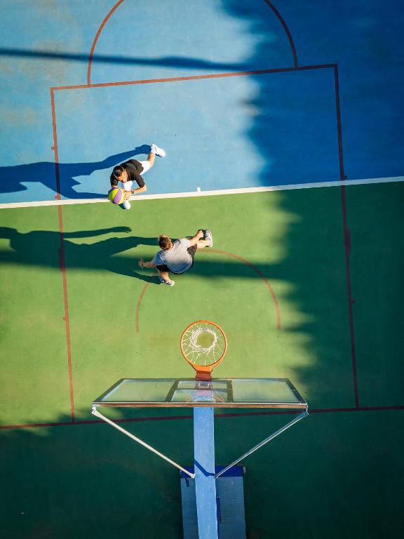 Divi spēlētāji spēlē basketbolu laukuma augšējā skatījumā, viens no viņiem tur bumbu, kamēr otrs gatavojas mest. Fons ir krāsainais basketbola laukums ar zilu un zaļu krāsu, kā arī grozs.