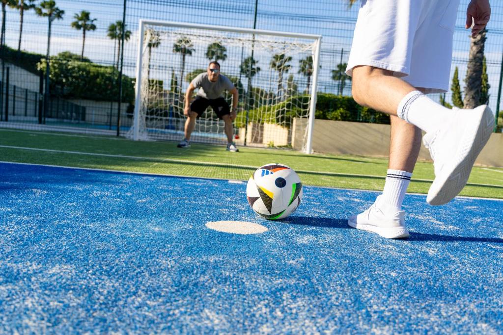Futbola laukums, kur spēlētājs izpilda sitienu uz vārtiem, kamēr vārtsargs gatavojas aizsargāt. Skaidra debesis un palmām apstādīts fons.