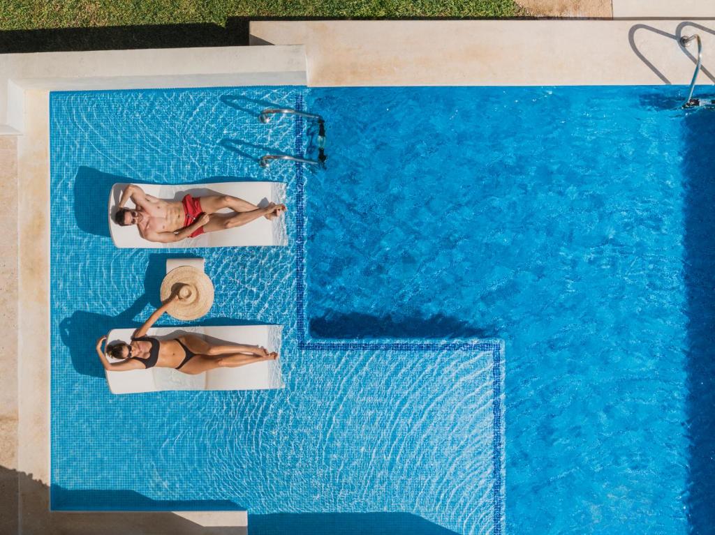 Divi cilvēki atpūšas uz pludmales krēsliem blakus peldbaseinam, baudot sauli un relaksāciju. Baseins ar dzidru, zilu ūdeni un zaļo zāli ap to rada mierīgu atmosfēru.