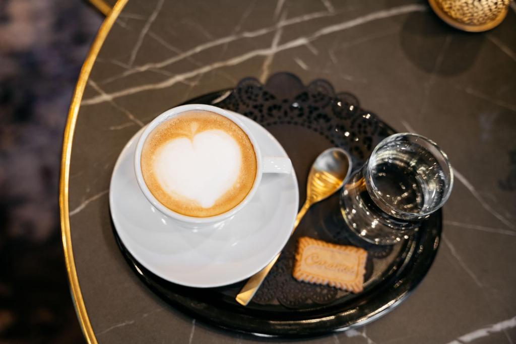 Kafijas krūze ar sirds formas putām uz balta šķīvīša, kopā ar ūdens glāzi un cepumiņu, pasniegta uz melna paplātes.