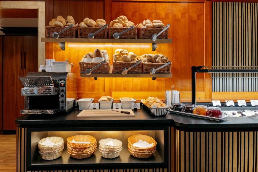 Brokastu stacija ar dažādu maizes izstrādājumu, ievārījumu un galda piederumu piedāvājumu, kas izvietoti modernā kafejnīcas vidē.