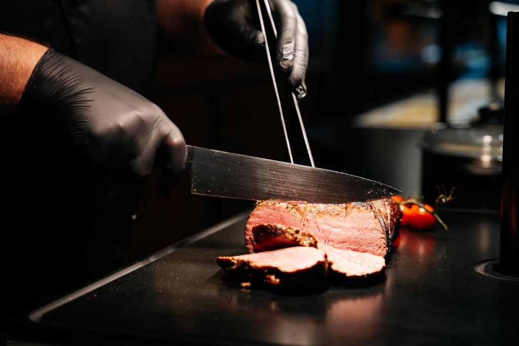 Šefpavārs ar melnām cimdiem sagriež sulīgu gaļu, izmantojot lielu nazi, blakus atrodas tomāti.