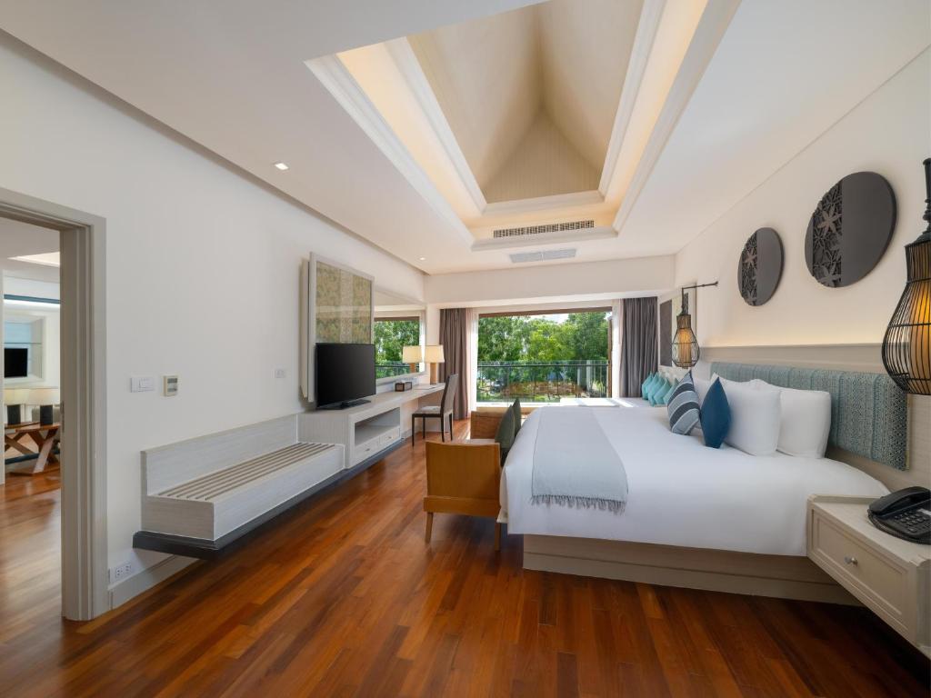 Mūsdienīgs guļamistabas interjers ar koka grīdu, plašu gultu un lielām logu durvīm, kas ved uz balkonu, piedāvājot dārza skatu. Telpā ir arī televizors un eleganti apgaismojumi.