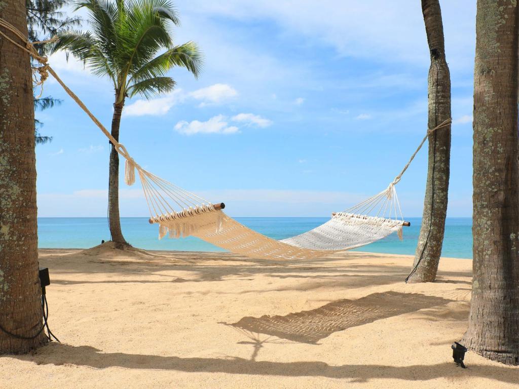 Atpūtas šūpuļtīkls, kas piestiprināts pie palmu kokiem, atrodas pludmalē ar skatu uz mierīgu okeānu un zilo debesu fonu.