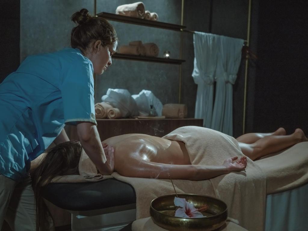 Masāžas sesija relaksējošā spa vidē, kur masieris koncentrējas uz klienta muguru. Gaisotne ir mierīga, ar dekoratīvām dvieļiem un ziediem.