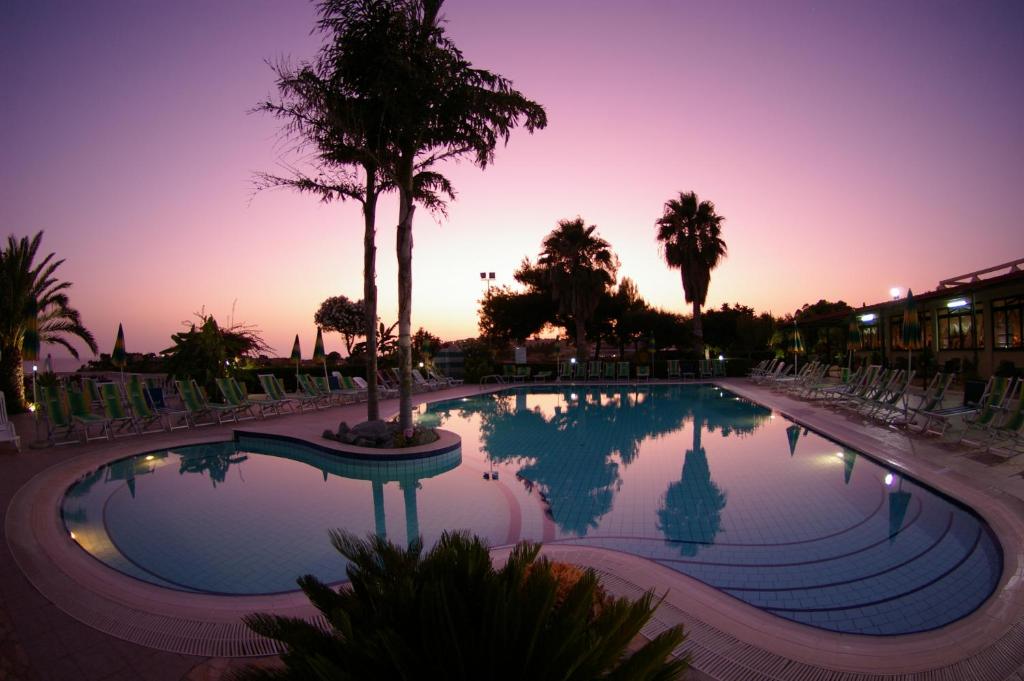 Peldbaseins ar palmām un saulrietu fona, radot mierīgu un relaksējošu atmosfēru. Šis attēls izceļ skaisto ainavu un viesnīcas peldbaseina dizainu.