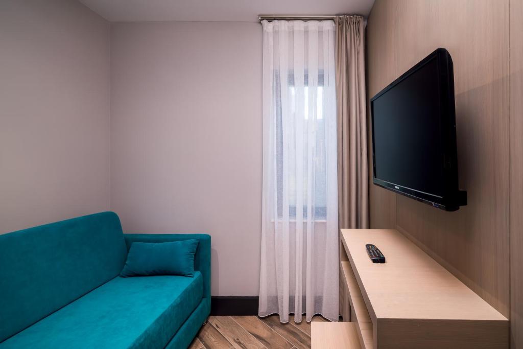 Mūsdienīga viesnīcas numura interjers ar zilu dīvānu, televizoru un logu ar caurspīdīgu aizkaru. Eleganta un vienkārša dizaina atmosfēra.