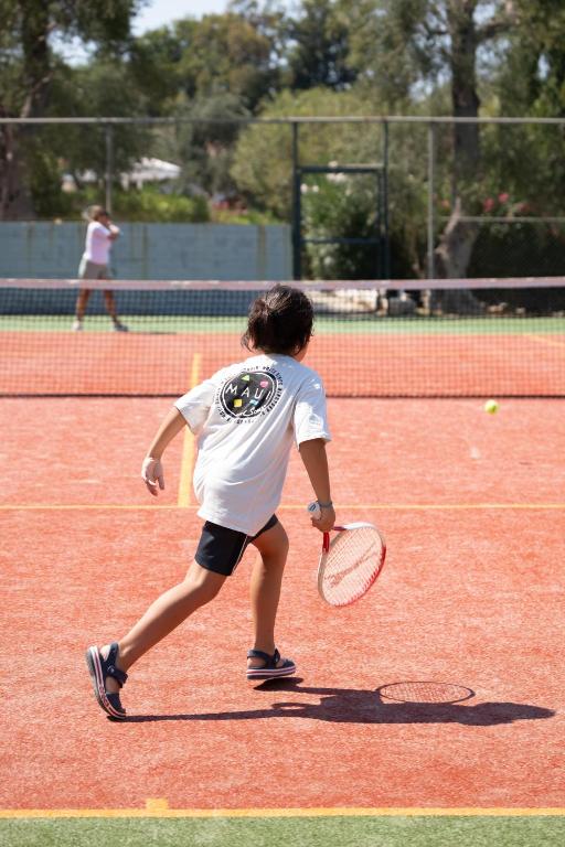Bērns skrien pa tenisa laukumu, turot raketi, kamēr fons ir redzams otra spēlētāja darbības. Tenisa laukums ir oranžs, un gaisma ir saulaina.