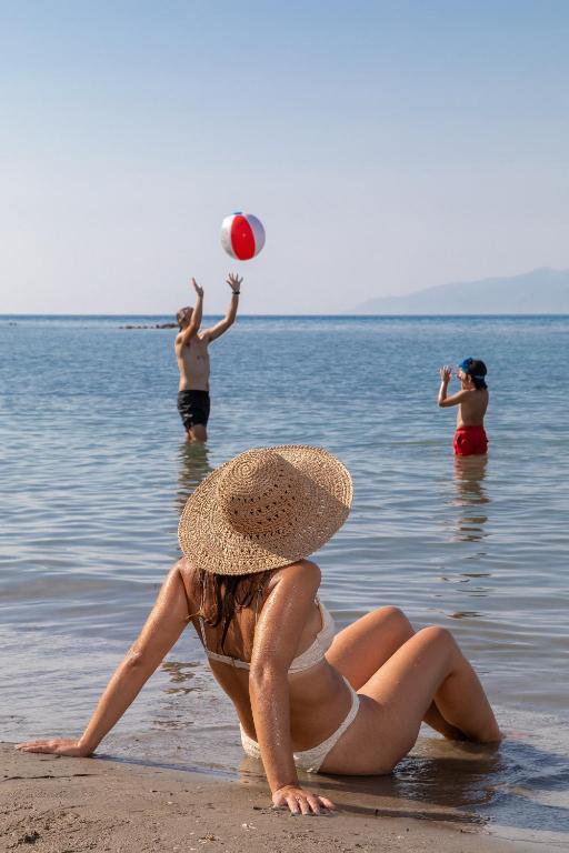 Pludmales aina ar cilvēkiem, kas spēlē ar pludmales bumbu ūdenī, bet sieviete ar saules cepuri atpūšas krastā.
