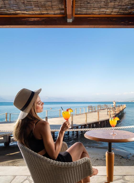 Sieviete ar cepuri sēž krēslā pie jūras, baudot kokteili, kamēr fons ir saulains skats uz laivu piestātni un mierīgu ūdeni.