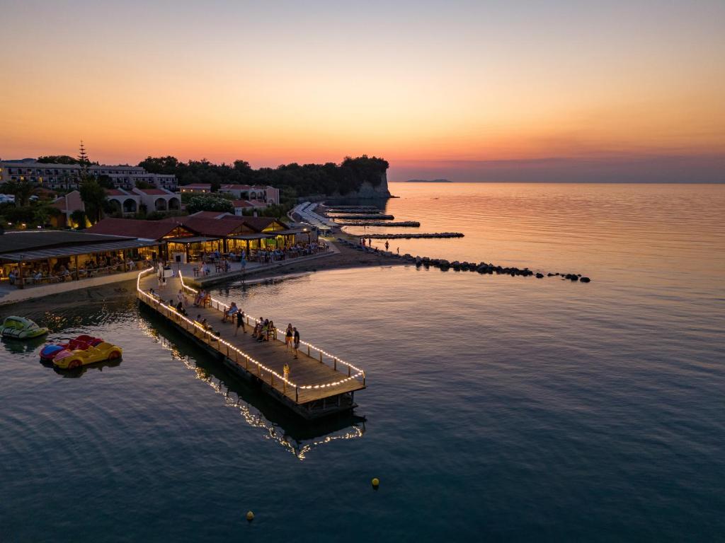 Saulrieta skats uz piekrasti ar promenādi un cilvēkiem, kas baudījuši vakara atmosfēru. Klusi viļņi un krāsains debesis piešķir mierīgu noskaņu.