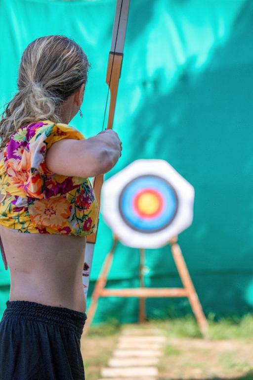 Sieviete šauj ar loku uz mērķi, kas ir izvietots zaļā fona priekšā. Viņas apģērbs ir krāsains ar ziedu rakstu, un fons ir saulains un skaidrs.