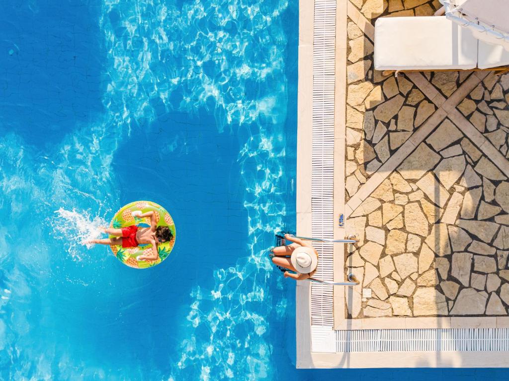 Bērns peld baseinā uz pludiņa ar ananāsu dizainu, kamēr pie baseina atpūšas pieaugušais ar saules cepuri. Ūdens ir dzidrs un mirdzošs, radot relaksējošu atmosfēru.