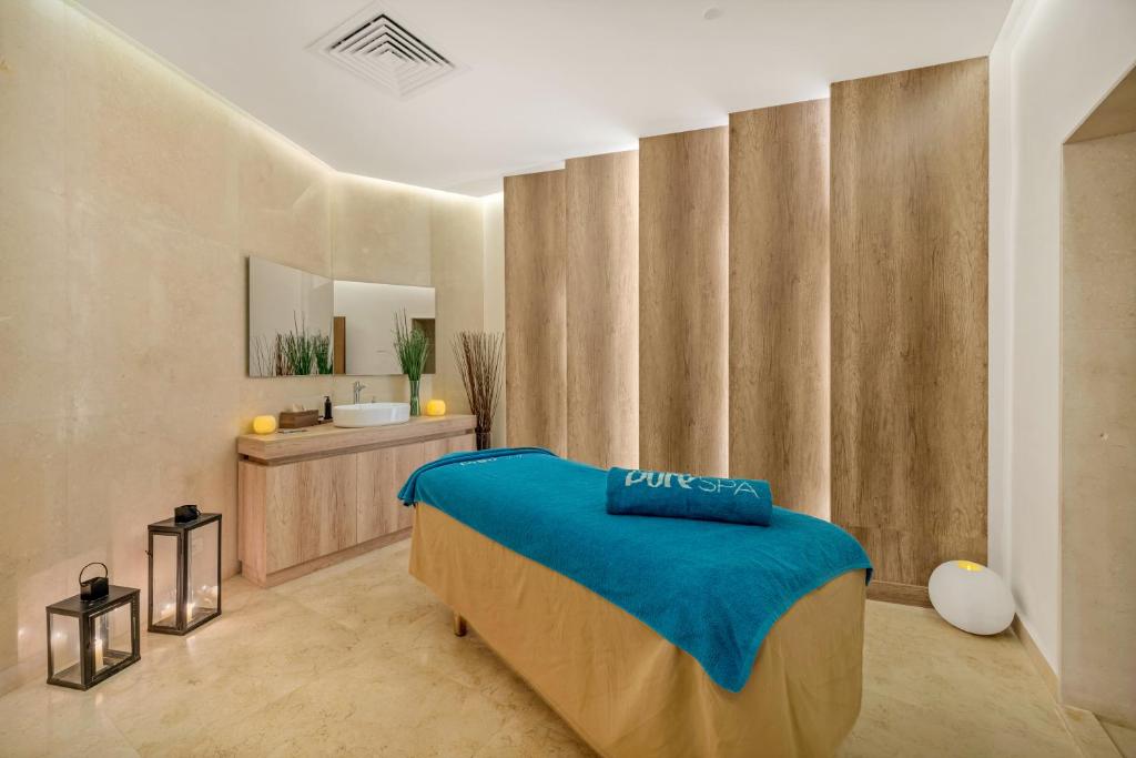 Mūsdienīgs spa telpas interjers ar masāžas galdu, kas klāts ar zilu dvieli, un elegantām dekorācijām. Telpā ir gaišas krāsas, koka akcenti un gaismas, kas rada mierīgu atmosfēru.
