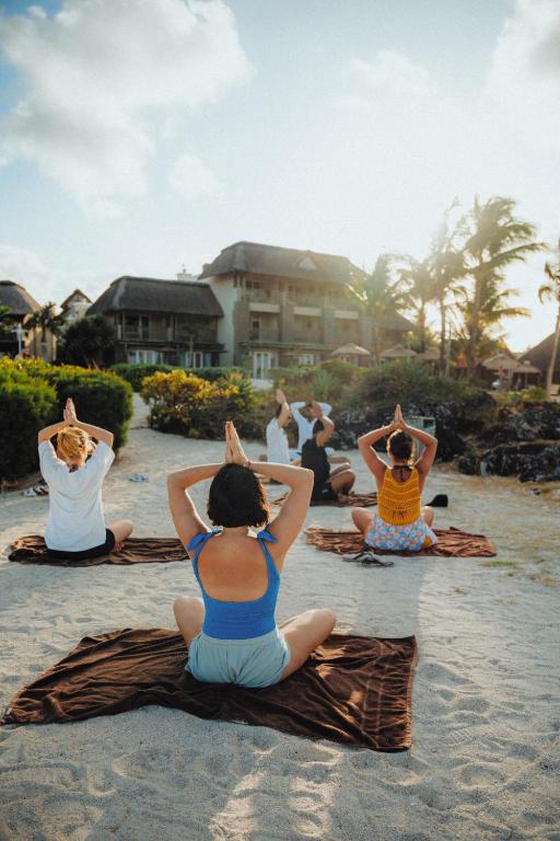 Grupas jogas nodarbība pludmalē ar dalībniekiem, kas veic meditācijas pozīcijas, baudot saulainu laiku un dabu.