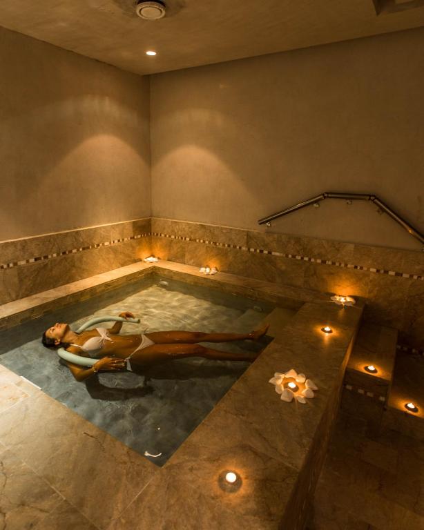 Relaksējoša spa vide ar cilvēku, kas guļ ūdenī, apgaismota ar svecēm un ziedlapiņām. Eleganti interjera elementi un maiga atmosfēra veido ideālu vietu relaksācijai.