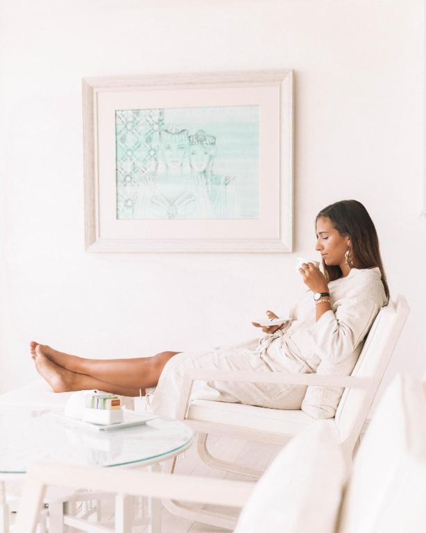 Sieviete, tērpta vieglā apģērbā, relaksējas baltā telpā, baudot dzērienu un skatoties uz telefonu. Fons ir dekorēts ar gleznu, kas piešķir atmosfēru.