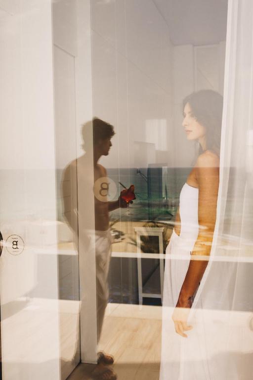 Attēls ar vīrieti un sievieti, kas atspoguļojas caur caurspīdīgu aizkaru, radot sapņainu atmosfēru. Fons ietver mierīgu jūras ainavu, piešķirot attēlam romantisku noskaņu.