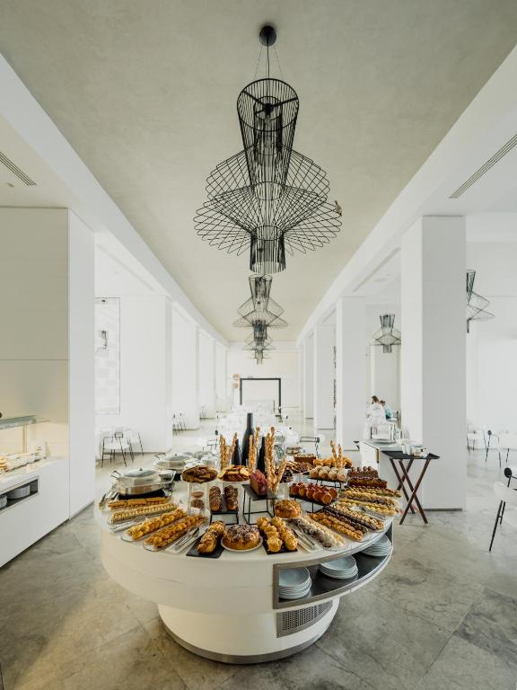 Elegants restorāns ar modernu interjeru, centrā apļa formā izvietota bufete ar dažādiem konditorejas izstrādājumiem un uzkodu šķīvjiem. Gaismas ķermeņi pie griestiem piešķir telpai īpašu šarmu.