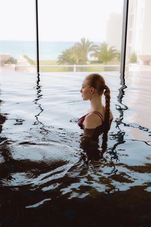 Sieviete ar pītiem matiem stāv ūdenī, skatoties uz jūru caur lieliem logiem, kas atspoguļo mierīgu ainavu.