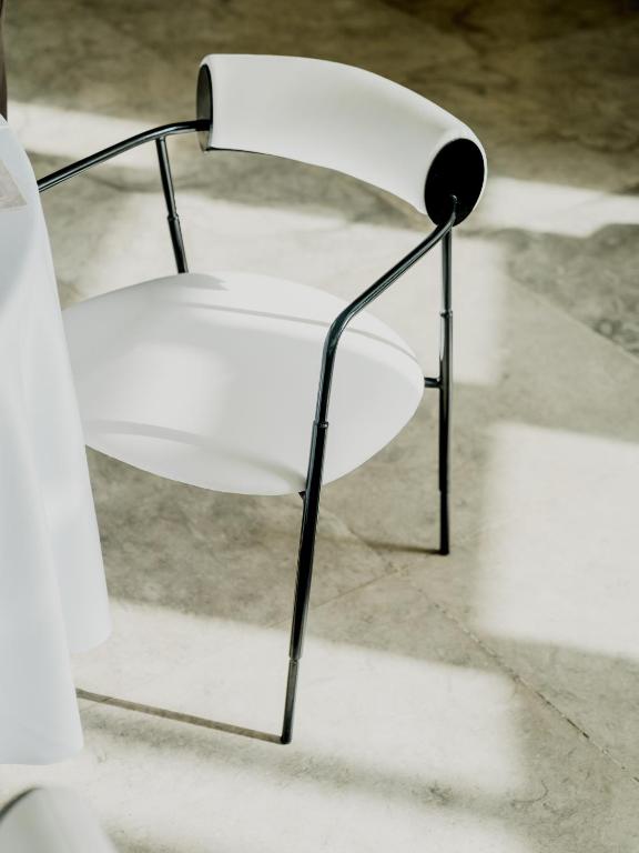 Mūsdienīgs balts krēsls ar melnu metāla rāmi, novietots uz gaismas piepildīta grīdas. Krēsla dizains ir vienkāršs un stilīgs, piemērots mūsdienu interjeram.