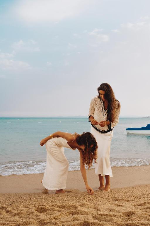 Divas sievietes pludmalē, viena no tām saliekusies, meklējot kaut ko smiltīs, otra stāvot blakus un skatoties uz viņu. Fons ir mierīgs jūras skats ar mīkstām viļņiem un skaidru debesu.