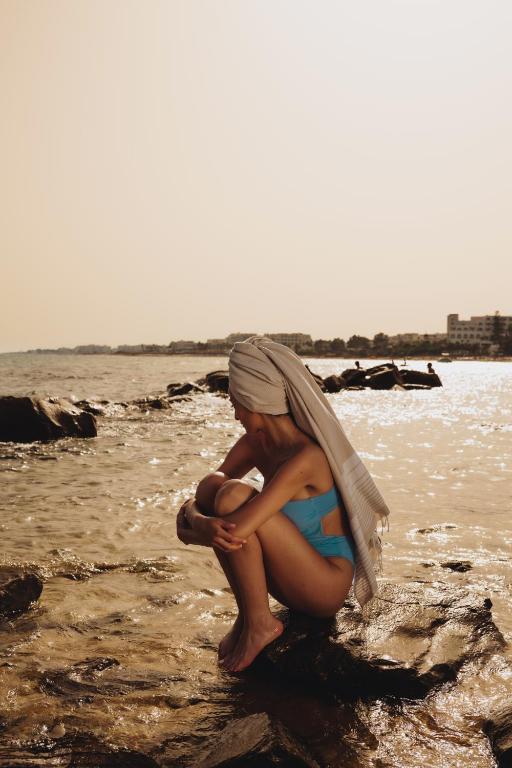 Sieviete ar dvieli galvā sēž uz klintīm pie jūras, tērpta zila peldkostīmā saulainā dienā. Fons ir mierīga jūra un pilsētas ainava.