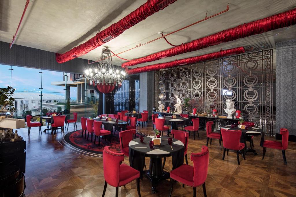 Elegants restorāna interjers ar sarkaniem krēsliem un greznu lustru. Lielie logi piedāvā skatu uz ārpusi, bet dekoratīvās statujas piešķir telpai māksliniecisku pieskārienu.