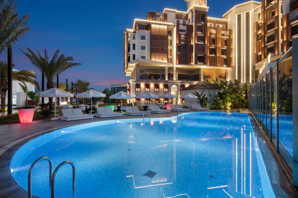 Mūsdienīgs viesnīcas baseins ar zilu ūdeni un saulessargiem, apgaismots vakara gaismā. Apkārt augstas palmas un eleganta arhitektūra.