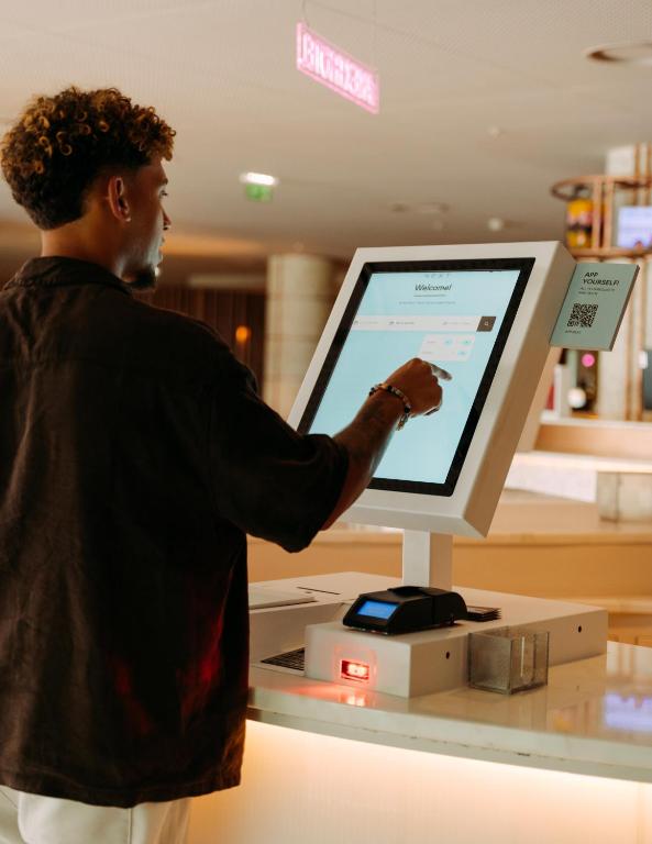 Jauns digitālais kiosks, kurā cilvēks izmanto skārienjūtīgu ekrānu, lai veiktu pasūtījumus vai reģistrētos. Moderns interjers un tehnoloģiskais risinājums, kas uzlabo apkalpošanu.