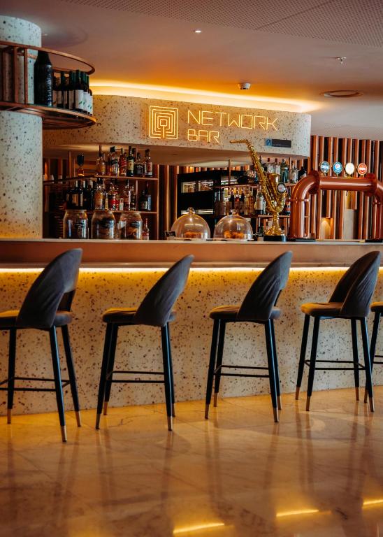 Mūsdienīgs bārs ar nosaukumu "NETWORK BAR", kas izceļas ar stilīgām krēsliem un labi aprīkotu bāra leju, apgaismots ar siltu gaismu.