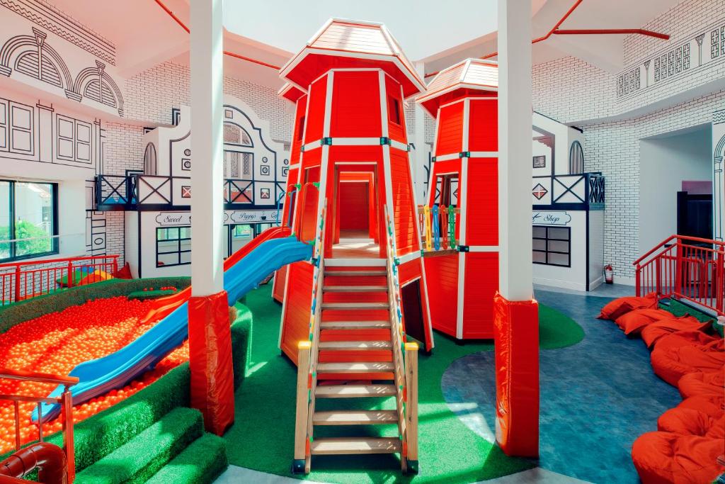 Krāsains bērnu rotaļu laukums ar divām sarkanām torņu struktūrām, slidkalniņu un bumbu baseinu. Interjers ir moderns un draudzīgs bērniem, ar mīkstām sēdvietām un zaļu zālāju.