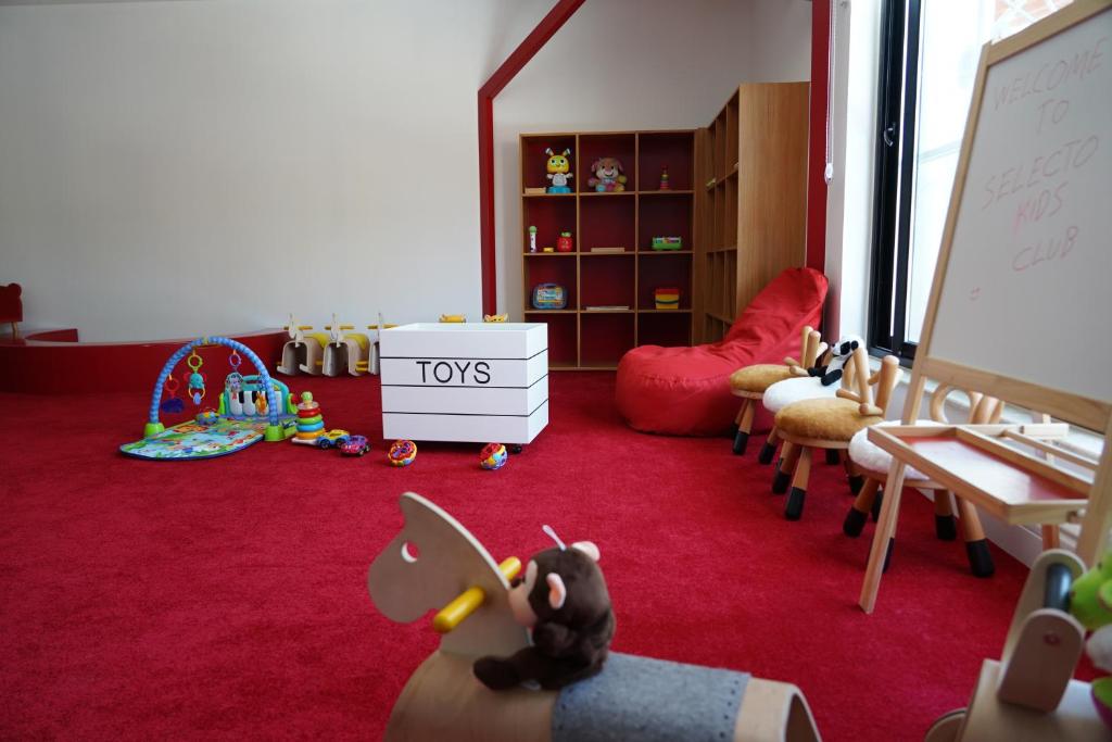 Bērnu istaba ar sarkanu paklāju, rotaļlietām un mīkstajām mēbelēm, kas nodrošina drošu un jautru vidi bērniem. Uz galda ir uzraksts 