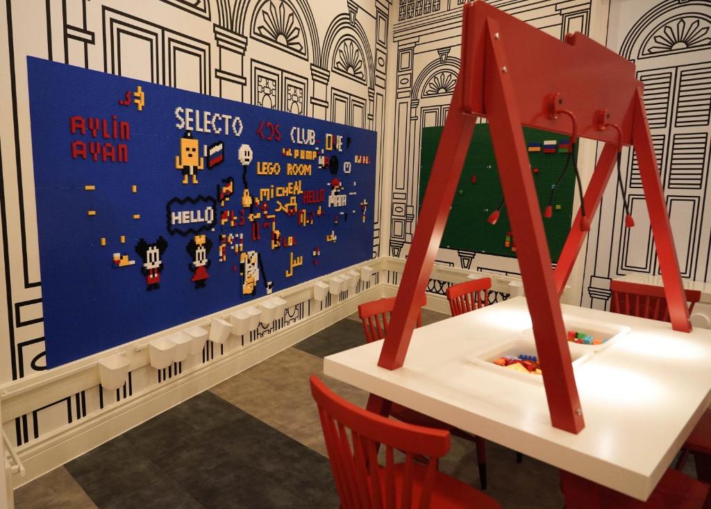 Rūpīgi izstrādāta Lego istaba, kurā redzams zils Lego panelis ar krāsainām figūrām un uzrakstiem, kā arī balta galds ar sarkaniem krēsliem.