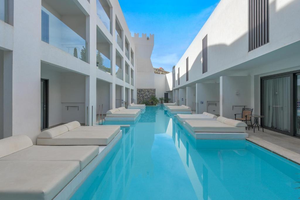 Moderns viesnīcas baseins ar saulessargiem un atpūtas zonām, kas atrodas starp baltām ēkām. Skats uz skaidru, zilu ūdeni un debesīm.