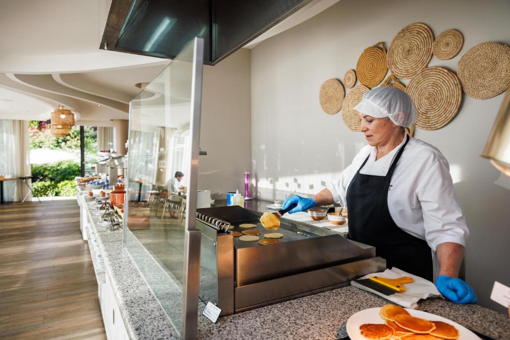 Konditorejas darbiniece gatavo pankūkas modernā virtuvē, izmantojot profesionālo aprīkojumu, kamēr fons rotājas ar dabīgu apgaismojumu un dekoratīvām detaļām.