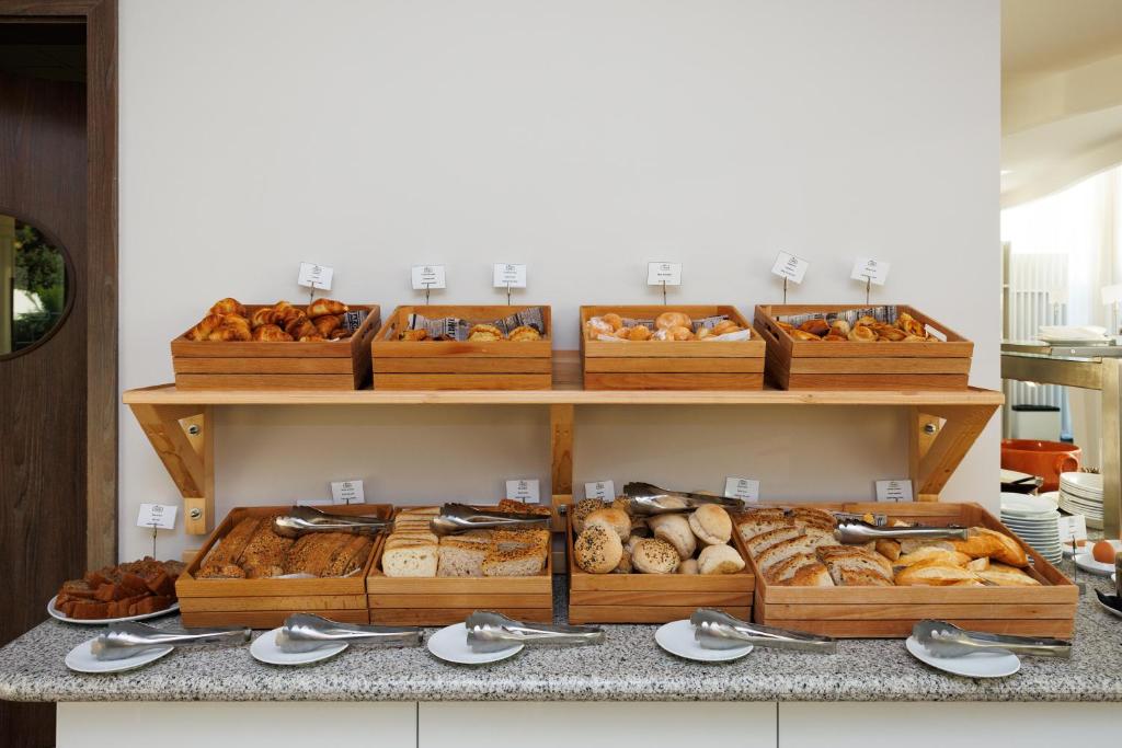 Brokastu bufete ar dažādiem maizes izstrādājumiem un konditorejas izstrādājumiem, sakārtota koka plauktā ar metāla karotēm un šķīvīšiem.