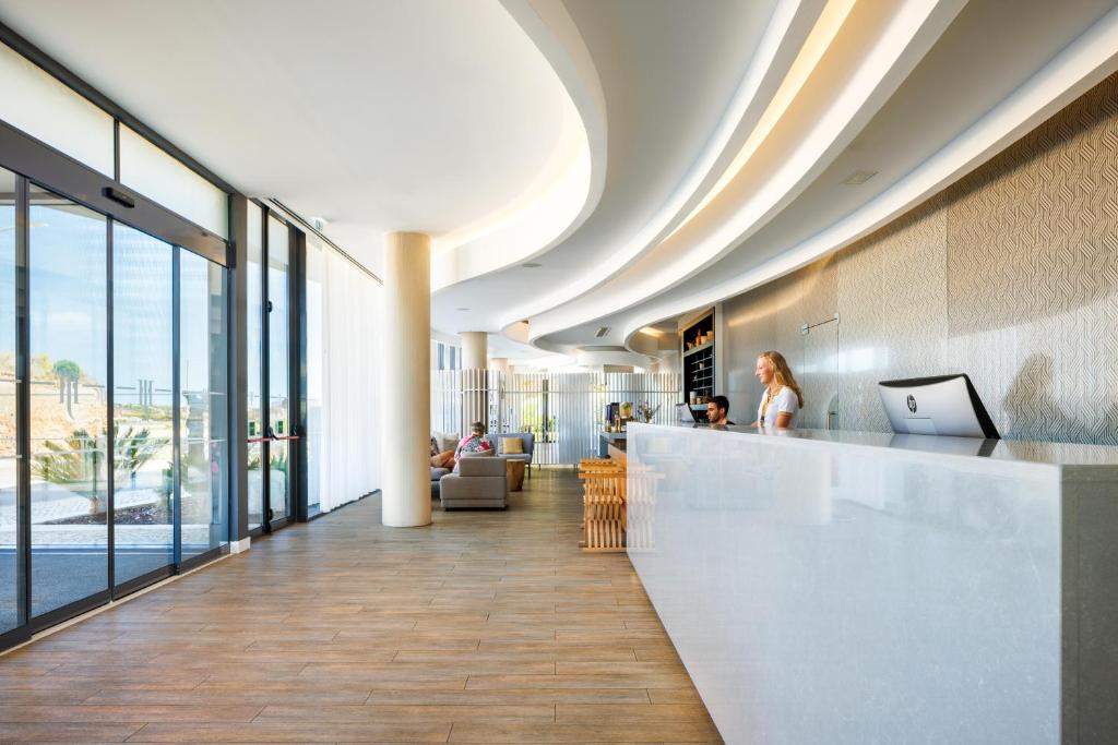 Mūsdienīgs viesnīcas reģistratūras interjers ar gaišiem toņiem un elegantām līnijām, kurā redzami viesi un darbinieki. Gaismas plūst caur lielajiem logiem, radot siltu atmosfēru.