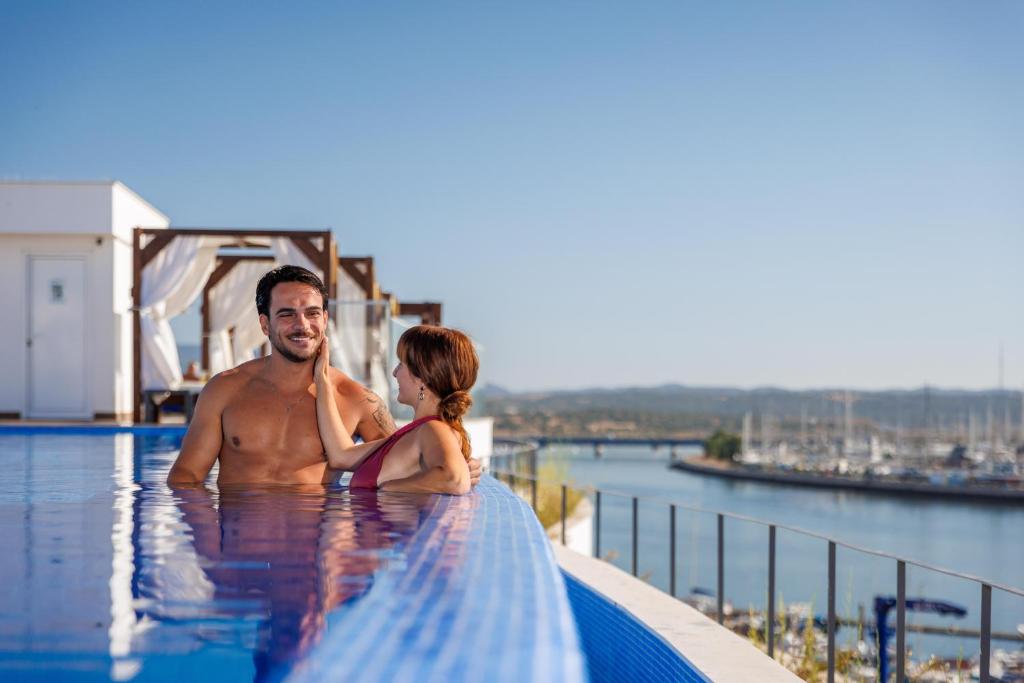 Pāris atpūšas bezgalības baseinā ar skatu uz ostu, baudot saulainu dienu un romantisku atmosfēru.