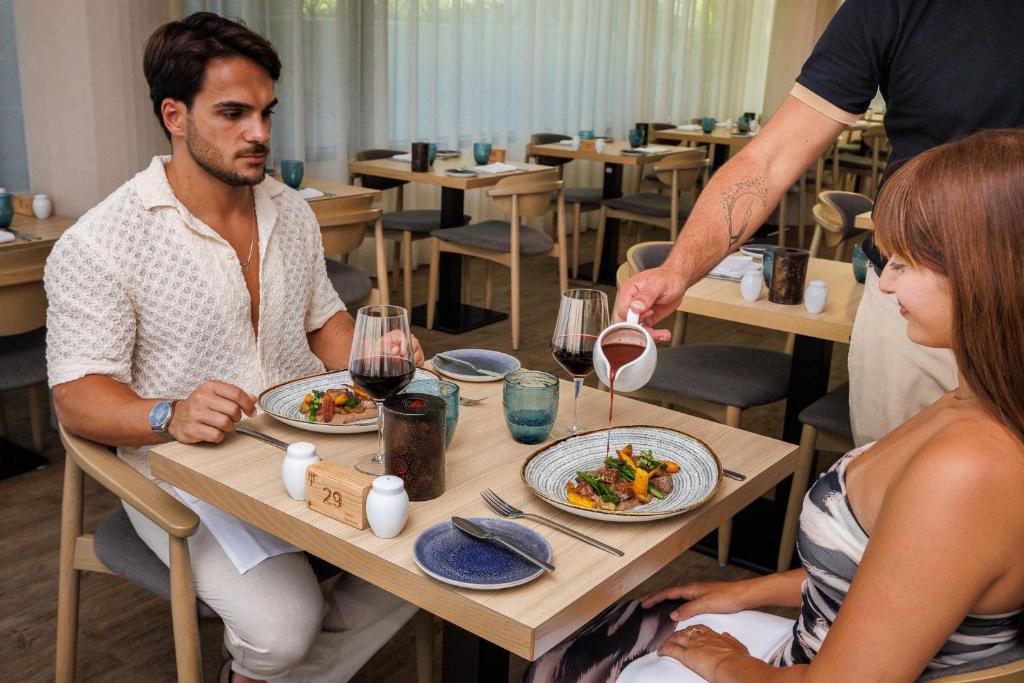Elegants restorāna brīdis, kur viesmīlis pasniedz ēdienu, kamēr pāris ar sajūsmu gaida maltīti. Galda dekorācija un atmosfēra rada patīkamu vakara noskaņu.
