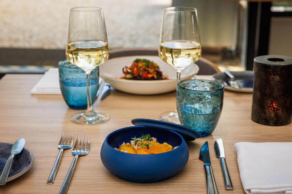 Elegantā galda iestatījums ar baltvīnu un krāsainiem ēdieniem, kas pasniegti stilīgās traukos. Mūsdienīga atmosfēra, kas aicina uz gastronomisku pieredzi.