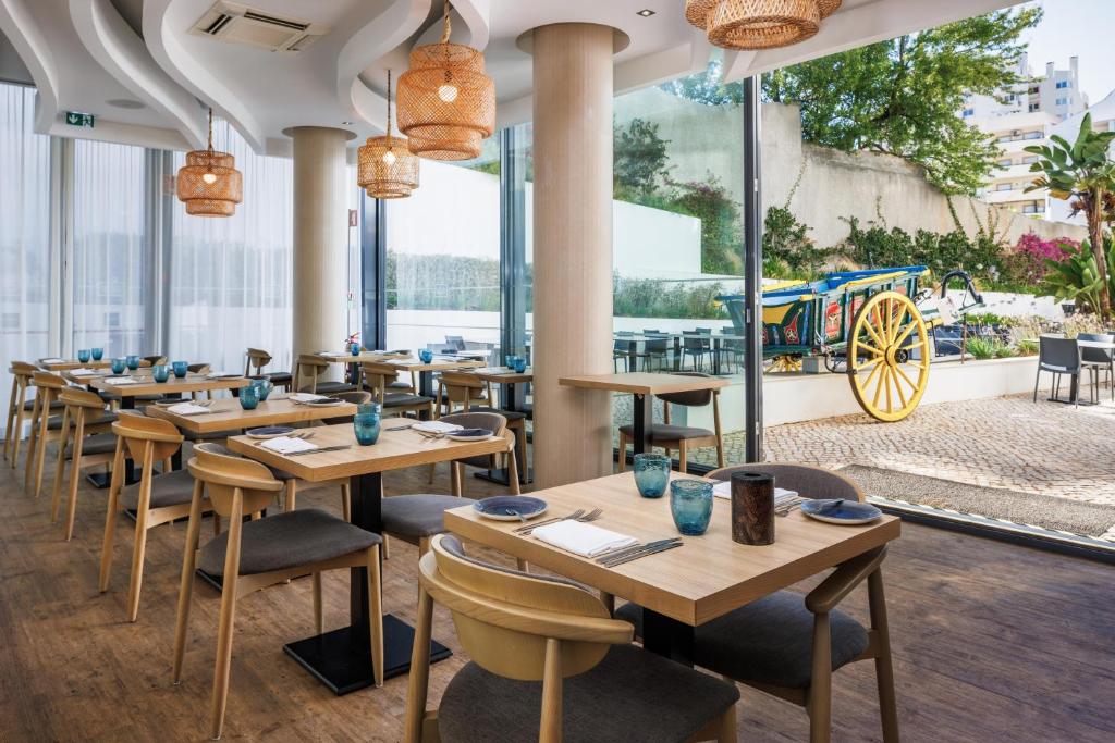 Mūsdienīgs restorāna interjers ar koka galdiem, stikla logiem un rotājošām lampām, kas sniedz patīkamu gaisotni. Skats uz ārpusi, kur redzama krāsaina ratiņa un zaļumi.