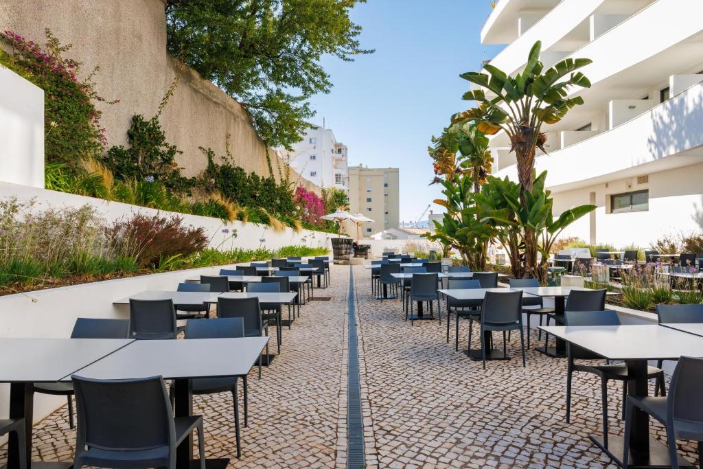 Āra kafejnīca ar melniem galdiem un krēsliem, kas izvietoti uz akmeņainas ietves, apkārt augošiem augiem un ziediem. Saulaina diena ar skaidru debesu fonu.