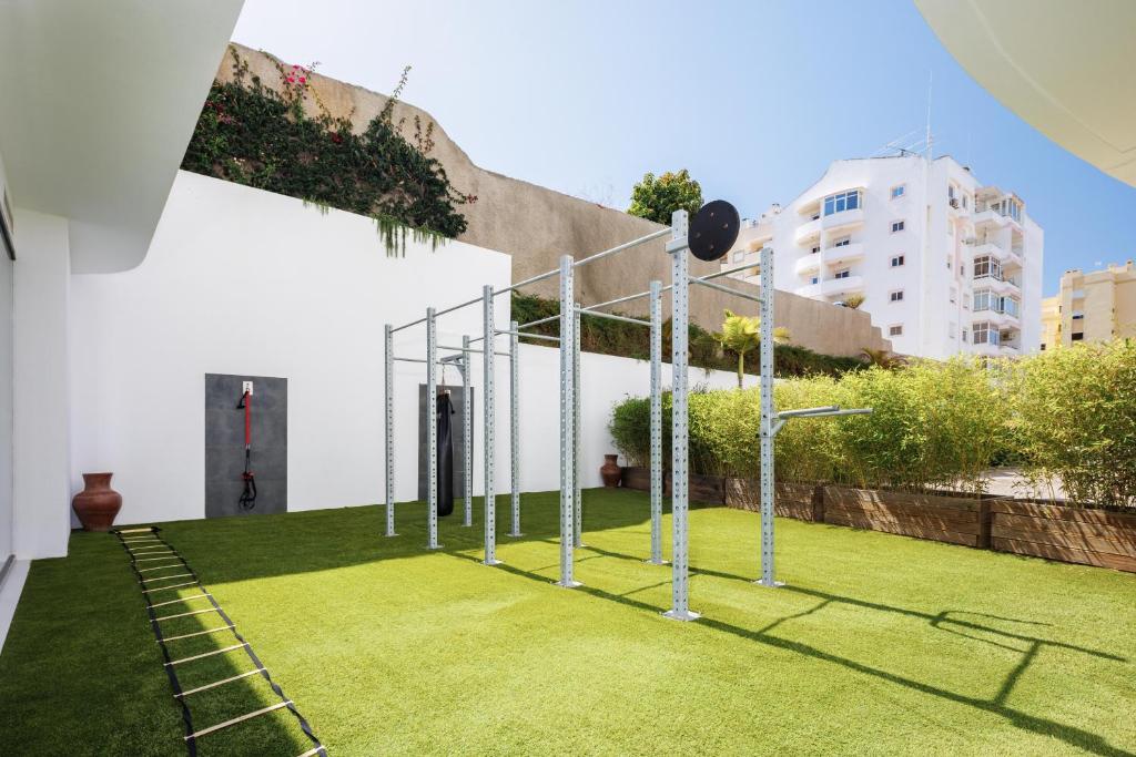 Āra trenažieru zona ar mākslīgo zāli un sporta aprīkojumu, ieskaitot svaru stieni un treniņu kāpnes. Fons veido pilsētas ainava ar zālienu un zaļumiem.