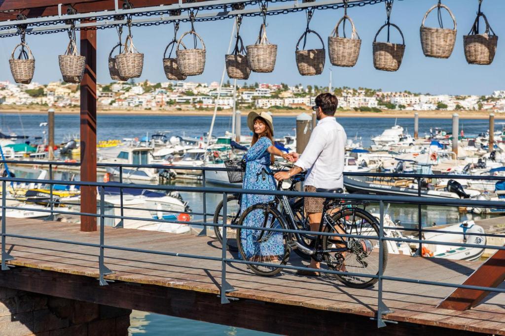 Pāris izklaidējas pie piestātnes, baudot saulainu dienu, ar velosipēdiem un laivām fonā. Sienas ir dekorētas ar groziem, kas piekārti pie pārsega.