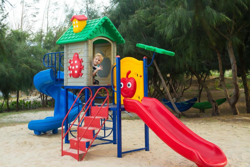 Bērnu rotaļu māja ar slide un kāpnēm, kurā bērns priecīgi iznāk no loga. Apstādījumi ar kokiem un smilšu laukums fonā.