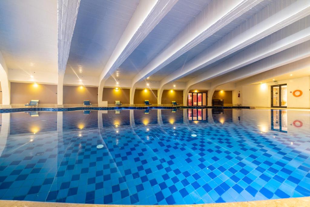 Moderns iekštelpu baseins ar zilām flīzēm un plašu telpu, kas apgaismota ar siltām gaismām. Atpūtas krēsli pie baseina rada relaksējošu atmosfēru.