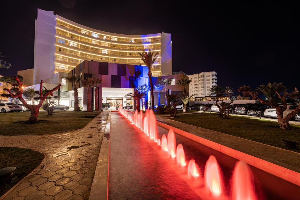 Mūsdienīgs viesnīcas eksterjers ar krāsainu apgaismojumu naktī un strūklaku, kas izstaro sarkanus ūdens plūsmus, rotājot zaļo zālienu un palmas priekšā.