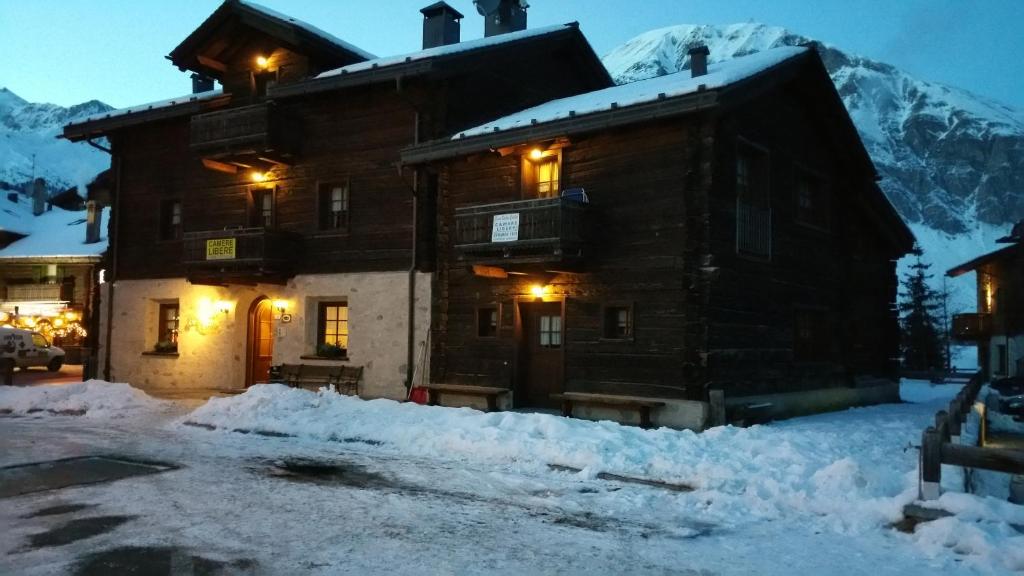 Koka māja ziemas ainavā, apgaismota ar siltām gaismām, apkārt klāta ar sniegu. Fons veido kalni, kas piešķir ainavai burvību.